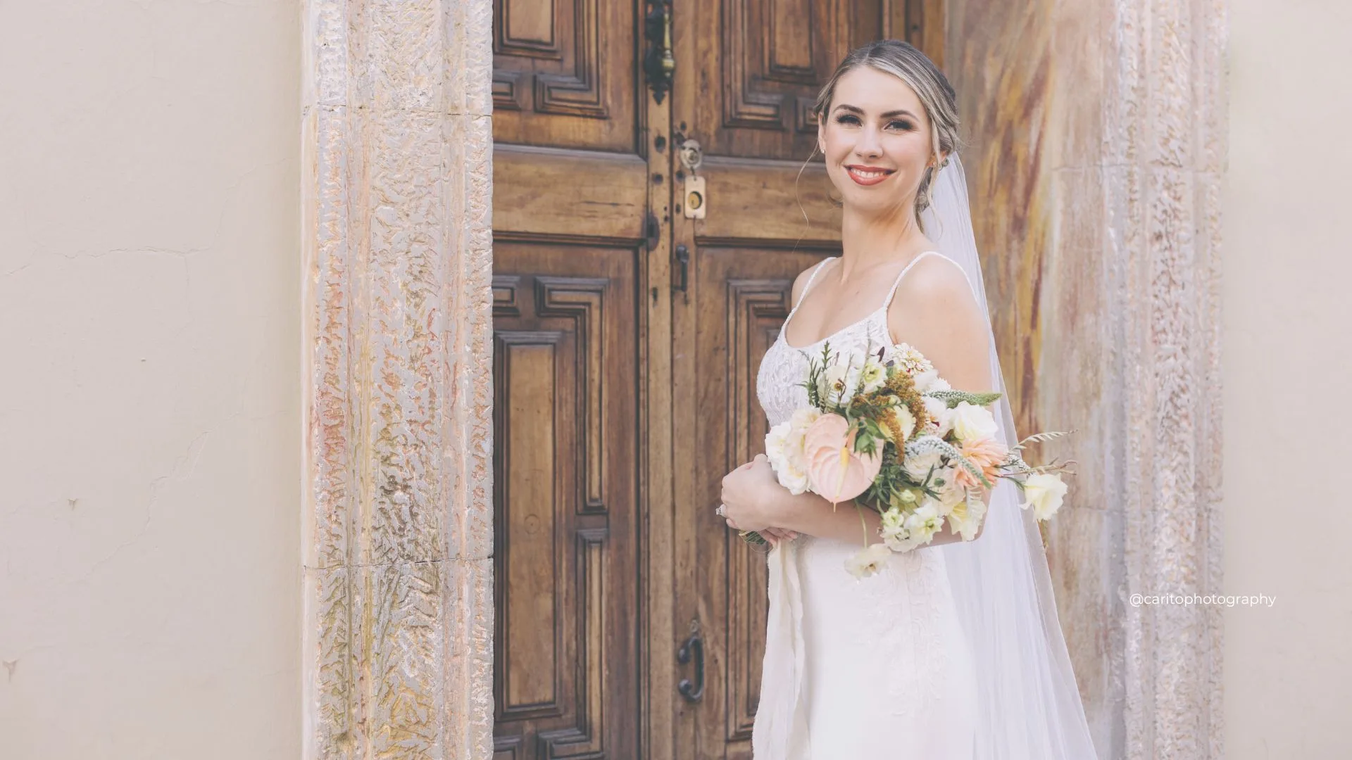 Bride with radiant makeup holding a pastel bouquet before carved wooden doors