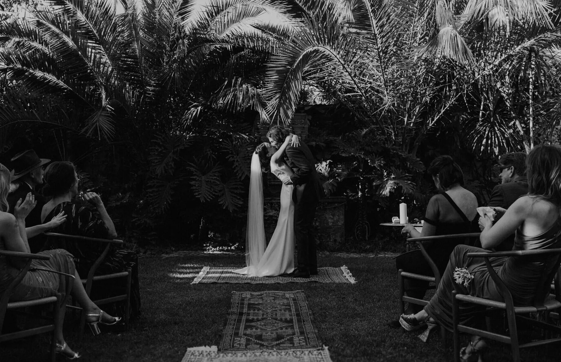 Black-and-white documentary photograph of a couple kissing during a garden ceremony