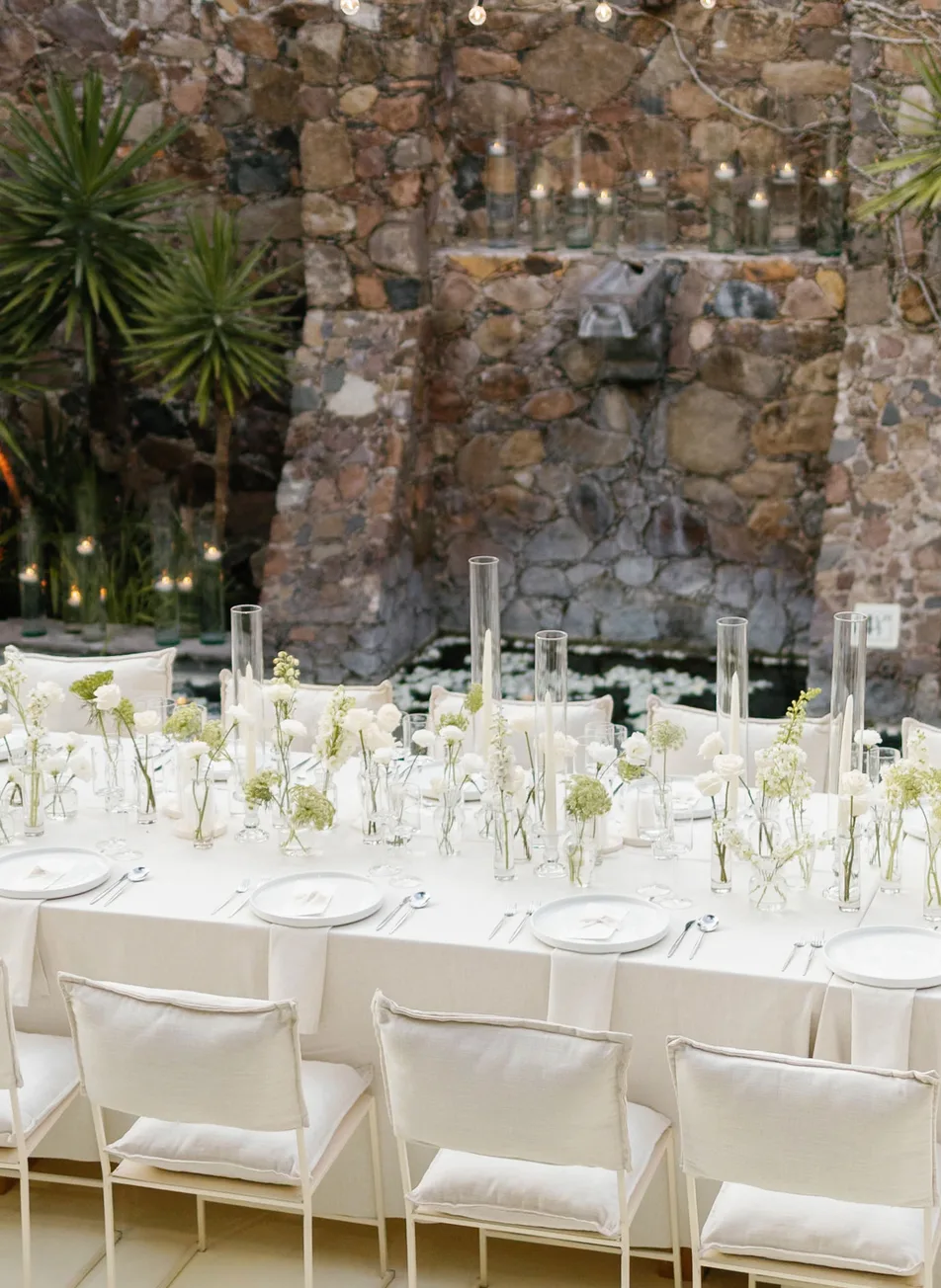 Elegant all-white reception table beside a stone fountain and floating candles