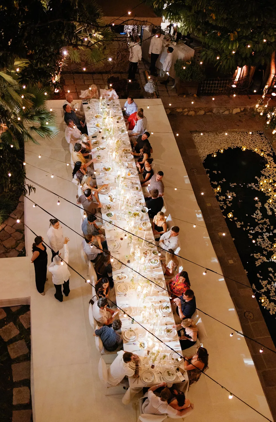 Overhead view of a candlelit banquet table beside a petal-filled reflecting pool