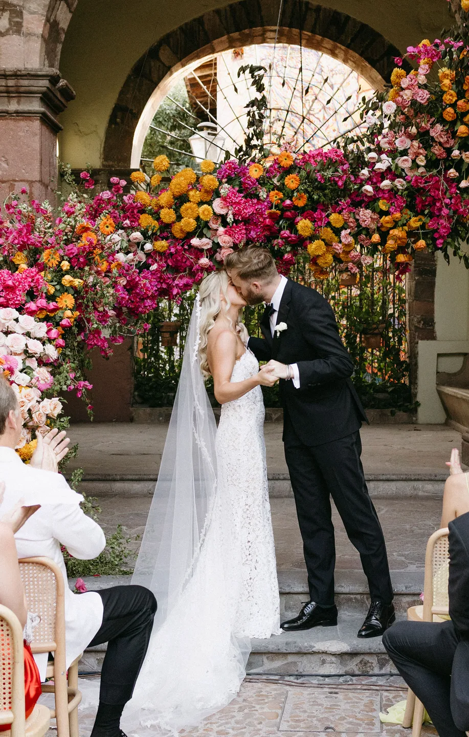 Newlyweds kissing beneath a dramatic colorful floral ceremony installation