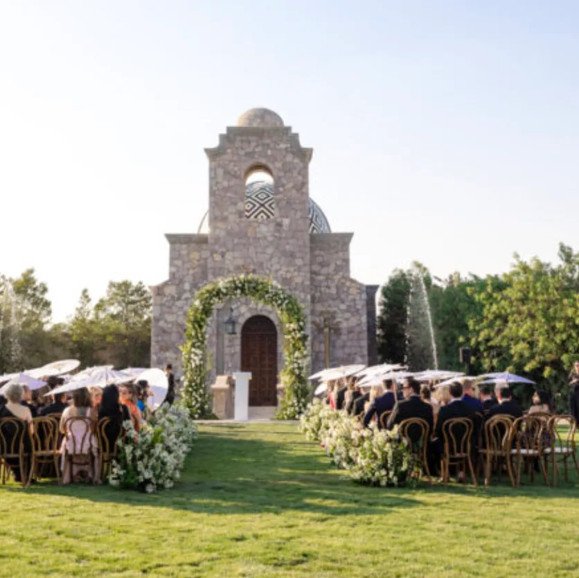 Outdoor wedding ceremony facing a stone chapel with a flower-lined aisle