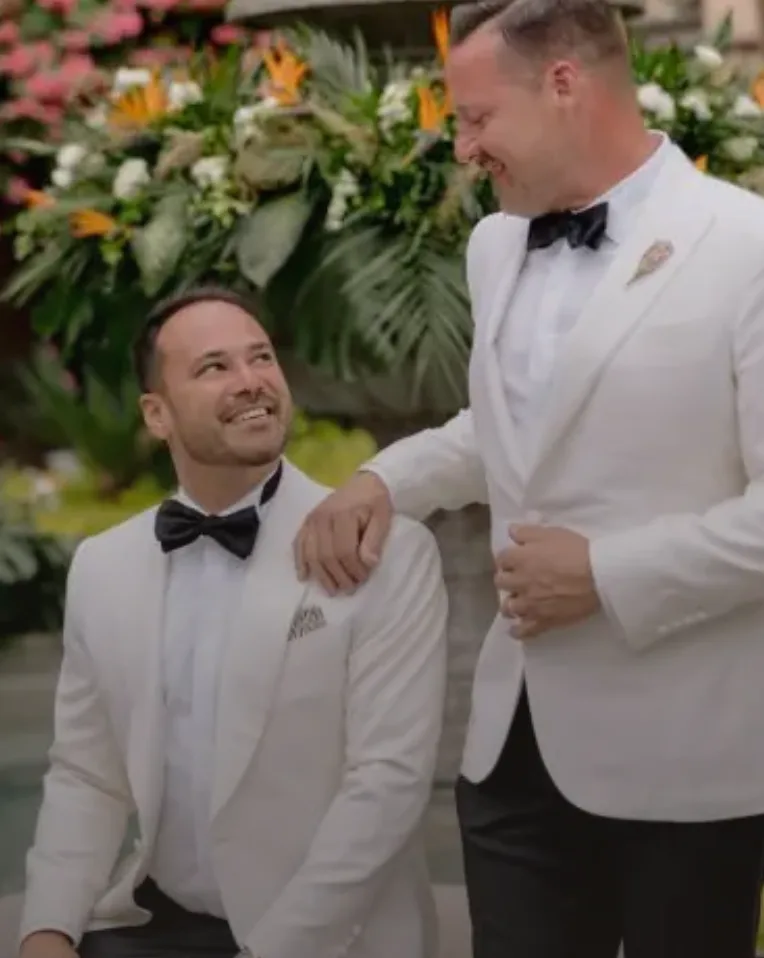 Two grooms smiling together before a tropical floral ceremony installation