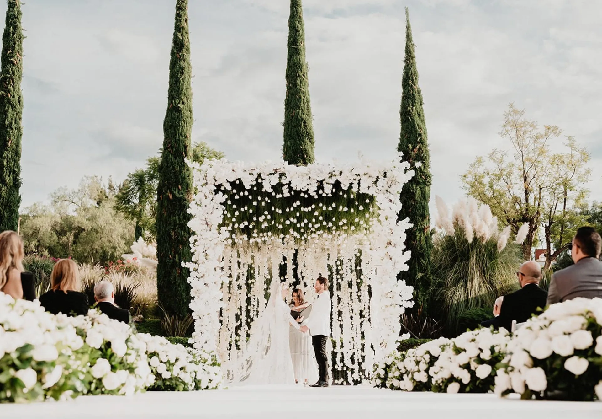 Outdoor wedding ceremony framed by cascading white flowers and tall cypress trees